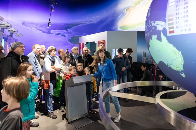 Eine Gruppe von Kindern und Erwachsenen nimmt in einem ineraktiven Ausstellungsst&uuml;ck an einer F&uuml;hrung teil.